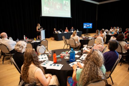 A group of people sitting at a table with a person standing in front of a screen.