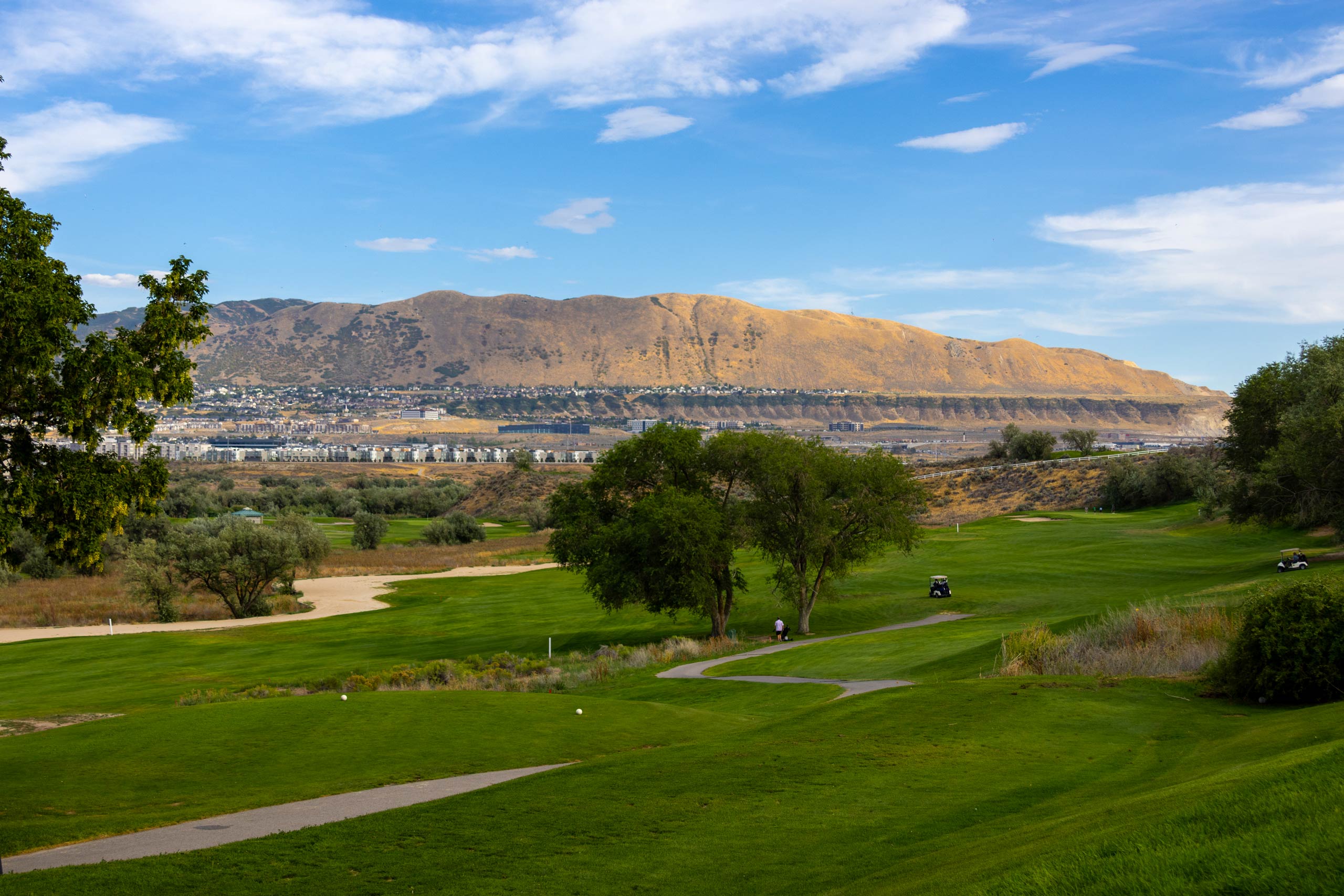 A fairway, golf cart path, and landscaping at Riverbend Golf Course in Riverton, Utah, with the city and mountains in the background.