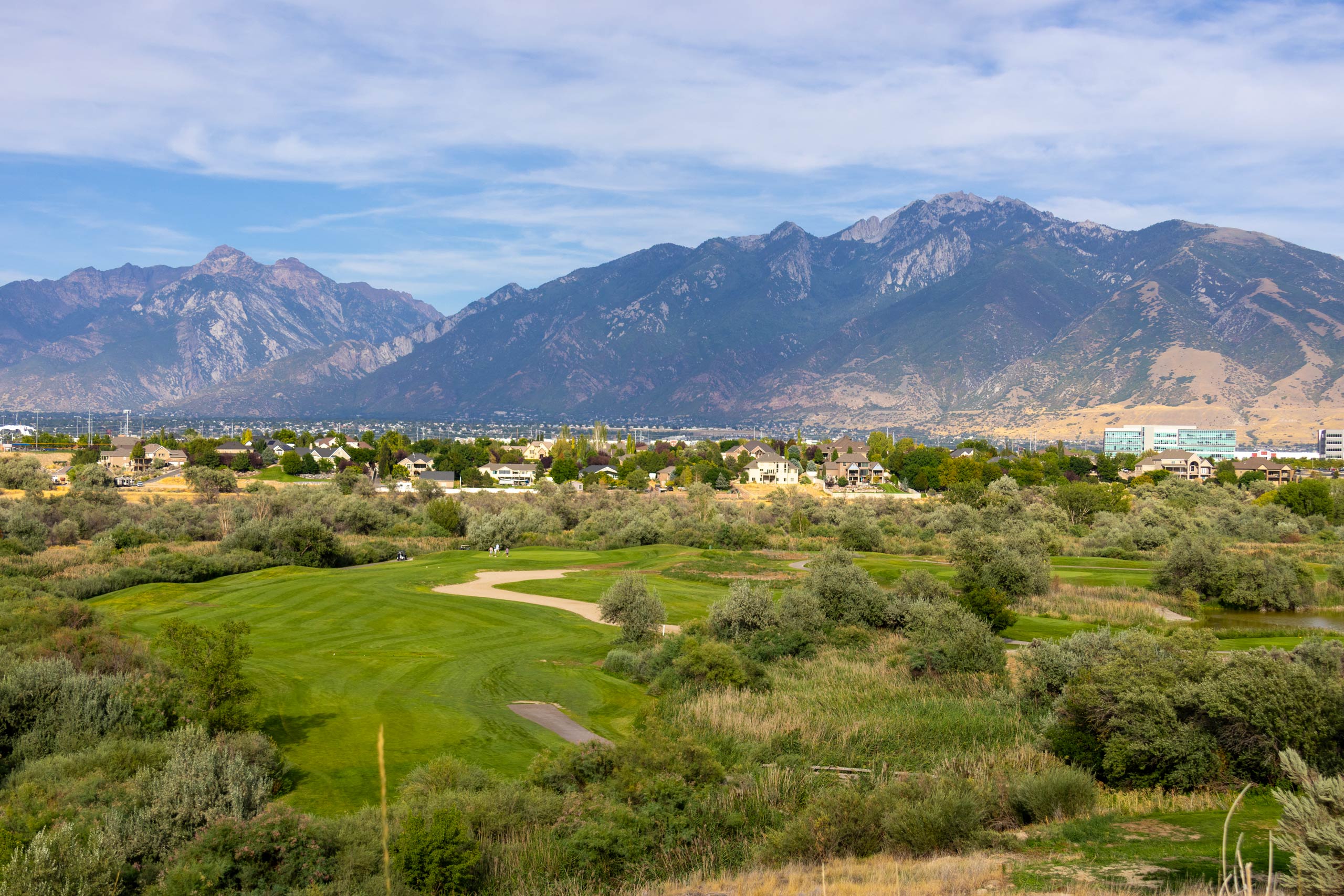 A broad view of a fairway and landscaping at Riverbend Golf Course in Riverton, Utah, with views of the city and mountains in the distance.